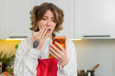Genç bir kadın, evde sağlıklı yemek yaparken mutfakta endişeli bir bakışla akıllı telefonu kontrol ediyor. Yetişkin kız kötü online mesaja tepki veriyor ve sağlıklı beslenmeye yemek hazırlarken diyet rutinini durduruyor.
