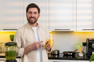 Orta yaşlı bir adam soyulmuş muzu işaret eder ve sağlıklı kahvaltı için mutfakta yeşil smoothie hazırlar. Genç adam sağlıklı beslenme hedefleri için evde meyve çerezi seçerek beslenme planını uyguluyor..