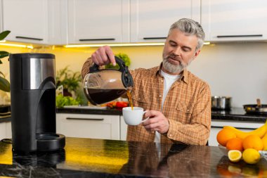 Orta yaşlı bir adam mutfaktaki damlatma makinesinden aromatik kahve döker. Günlük idmandan sonra gülümser. Olgun adam evde sıcak içeceğin tadını alıyor. Enerji odaklanma motivasyonu için sakin kahvaltı rutininin tadını çıkarıyor..