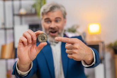 Merkez ofisteki orta yaşlı işadamı, kamera paylaşımındaki yatırım potansiyeline Bitcoin gösteriyor. Masadaki serbest çalışan adam kendine güvenen izleyicileri kripto para fırsatına davet ediyor..