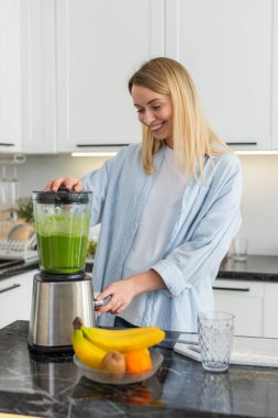 Orta yaşlı kadın smoothie 'yi elmalı ıspanaklı ıspanaklı spirulina klorellasıyla harmanlıyor sonra da ev mutfağında kahvaltı için bardağa döküyor. Genç kız sabah beslenmesi ve enerjisi için sallanıyor..