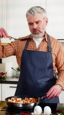 Orta yaşlı bir adam elektrikli soba baharatında sebze pişiriyor sonra da ev mutfağında sağlıklı beslenmek için yemek tadıyor. Yetişkin adam diyet yemeği hazırlarken gülümsüyor. Spor salonunda kilo kaybı yaşıyor.