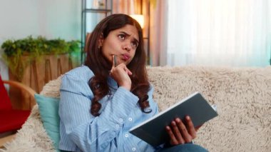 Hintli genç bir kadın, günlüğünü evde kanepede hafif bir gülümseme ve rahat bir duruşla yazıyormuş. Arap kızı güne barışçıl duygularla, kişisel zamanlarıyla ve içeride yumuşak iç huzuruyla yansıyor.