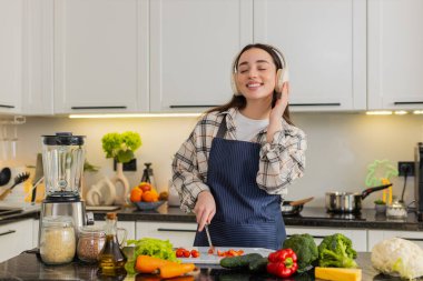 Genç bir kadın, temiz beslenme için evde vejetaryen salatası yapan kulaklık takan domatesleri kesiyor. Kız evde sakin bir şekilde sağlıklı yemek hazırlarken müziği seviyor..