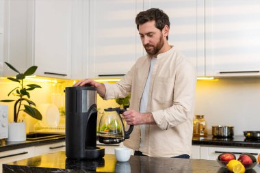 Orta yaşlı bir adam sıcak kahve koyar sonra zengin bir koku alır ve sabahın erken saatlerinde ev mutfağında gülümser. Genç adam beyaz bardaktan sıcak bir içecek yudumluyor. Sakin bir kahvaltı başlıyor ve kafein içeride artıyor..