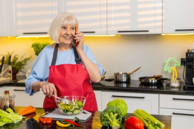 Yaşlı bir kadın, ev mutfağındaki telefon görüşmesi sırasında sağlıklı beslenme için taze sebze salatasını kasede karıştırıyor. Yaşlı büyükannem aile haberlerini paylaşıyor ve beslenme hazırlıklarını evde sakin bir şekilde yapıyor..