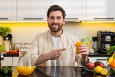 Orta yaşlı bir adam mutfaktaki bir bardak portakal suyunu göstererek gülümsüyor ve iyileşmek için vitamin içkisinin tadını çıkarıyor. Genç adam günlük enerji kondisyonu için sağlıklı beslenme seçimi ev yemeği rutinini teşvik ediyor..