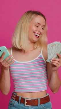 Akıllı telefonlu bir kadın tüm samimiyetiyle neşe dolu bir galibiyet, bir dolar nakit para alması, zaferi kutlaması piyango oyunu uygulaması kumarhane piyango ikramiyesi. Sarışın kız pembe arka planda izole edilmiş.