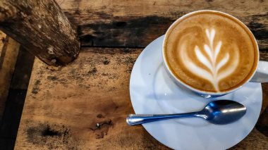 Cappuccino, sıcak ve davetkar bir atmosfer yaratmak için, rahat kafedeki ahşap masada kaşıkla beyaz çay tabağının üzerinde oturur..
