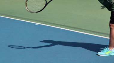 Tenis oyuncusu güneşli bir gün maçında açık havada topa servis yapıyor, atletizm gösteriyor ve rekabetçi ortama odaklanıyor..