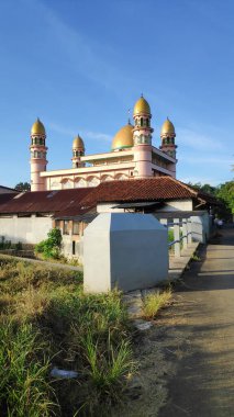 Cami, açık mavi gökyüzünün altında altın kubbelerle ayakta duruyor. Huzurlu bir mahallede İslami mimari ve ruhani huzur sergiliyor..
