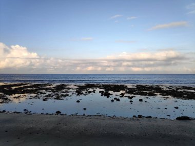 Plajda sakin bir yaz günbatımı. Altın güneş ışınları okyanus dalgalarını ve kumlu sahili yansıtıyor. Mavi gökyüzü bulutlarla ve uzak kuşlarla dolu. Gök mavisi titreşim