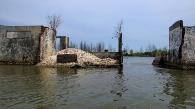 Sığ sularda terk edilmiş beton duvarlar, kabuk yığınları ve seyrek ağaçlarla çevrili, kıyı yerleşimlerinin yavaş çöküşünü ve çevresel kırılganlığı sembolize ediyor.