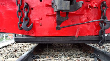 Tampon ışını, bağlantı kancasını, zincirleri ve sinyal ışıklarını gösteren eski bir kırmızı tren lokomotifinin ayrıntılı görüntüsü çakıl raylarında dinleniyor, gücü, dayanıklılığı ve geleneksel demiryolunu temsil ediyor.