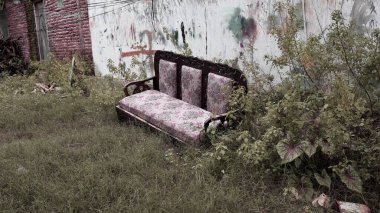 Çimenli zemine bırakılmış eski ahşap yastıklı eski bir kanepe, eskimiş grafiti ve sürünen bitkilerle kaplı yıpranmış bir duvara yaslanmış, kentsel bir bozunma atmosferi yaratıyor..