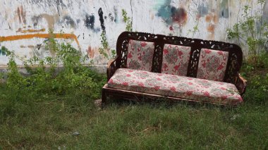 Yıpranmış ahşap bir kanepe, uzun çimenlerin üzerinde terk edilmiş, duvarın arkasında, üzerinde grafiti izleri ve yabani bitkiler olan, ihmal ve kentsel terk edilmeyi yansıtan, kırık beton bir duvar ile duruyor.