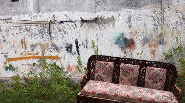 Terk edilmiş, kırmızı desenli döşemeleri olan terk edilmiş ahşap bir kanepe çimenli zeminin üzerinde, duvarda grafiti ile işaretlenmiş, güçlü bir terk edilmişlik ve çürüme hissi yakalıyor..