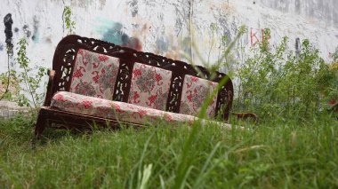 Yastıkları yıpranmış klasik bir açık kanepe çimenlerin üzerinde duruyor, solmuş grafiti ve sürünen bitkilerle çevrili kirli bir duvar tarafından çerçevelenmiş, şehir ihmalkarlığını ifade ediyor..