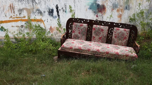 Yıpranmış ahşap bir kanepe, uzun çimenlerin üzerinde terk edilmiş, duvarın arkasında, üzerinde grafiti izleri ve yabani bitkiler olan, ihmal ve kentsel terk edilmeyi yansıtan, kırık beton bir duvar ile duruyor.