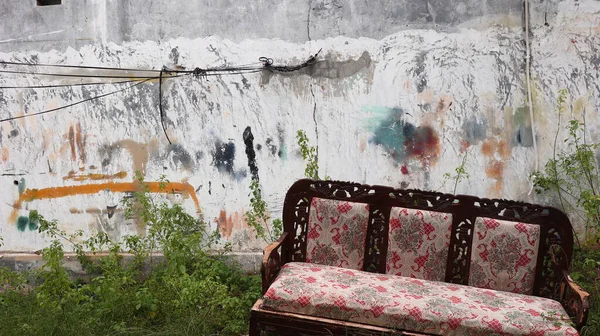 Terk edilmiş, kırmızı desenli döşemeleri olan terk edilmiş ahşap bir kanepe çimenli zeminin üzerinde, duvarda grafiti ile işaretlenmiş, güçlü bir terk edilmişlik ve çürüme hissi yakalıyor..