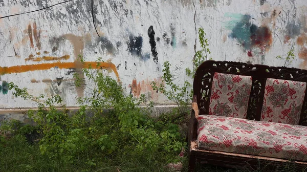 Eski bir kanepe, çimen ve yabani otlarla çevrili, solmuş graffitilerle lekeli bir duvarın önünde duruyor, ihmal, zaman ve kentsel bozulmayı gösteriyor..