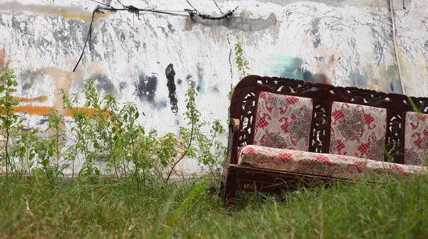 Kırmızı döşemeli, unutulmuş ahşap bir kanepe. Çimlerin üzerinde, duvarın yanında terk edilmiş. Kayıp, çürüme ve ihmal hikayelerini çağrıştırıyor..