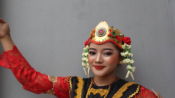 Young girl wearing a vibrant red and gold traditional costume poses gracefully against a plain wall, showcasing elegant makeup, ornate headpiece, and cultural heritage, Pekalongan November 6, 2025