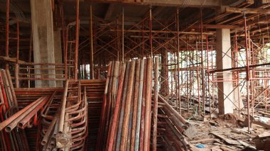 İnşaat halindeki bir binanın paslı iç manzarası, karmaşık paslı metal iskeleler, sağlam beton sütunlar ve dağınık ahşap döküntüler, gelişimin ilk aşamalarını vurguluyor..