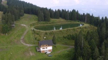 Slovenya 'nın Kope kentinde çam ormanlarıyla çevrili gölet ve dağlarda bir ev, Arial solda daireler çiziyor.