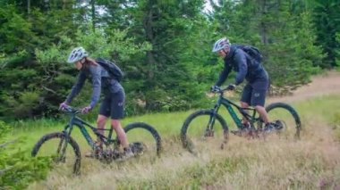 Etrafı çam ağaçlarıyla çevrili yolda giden iki yetenekli MTB motorcusu. Yavaş çekim
