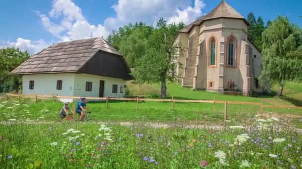 花の牧草地を持つ古い教会を通過する2人の自転車 — ストック動画