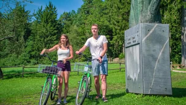 公園の記念碑の前を自転車で歩く若いカップルが景色を楽しむ — ストック動画
