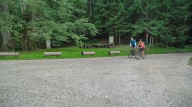 Dağ bisikleti süren atletik çift, ormanın önündeki asfalt yolda giderken büyük yaşlı bir ağacı işaret ediyor. İzleme görüntüsü.