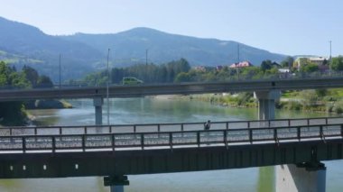 Adam, Drava nehri üzerindeki yaya köprüsü boyunca yürür. Elinde telefonu, havada uçan insansız hava aracı görüntüsüyle.