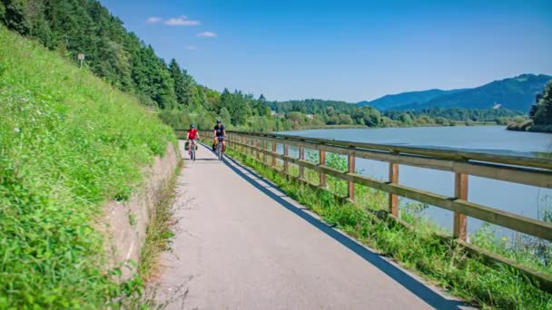 緑豊かな風景の中 湖の端の道を自転車で走るカジュアルカップル スローモーションショット — ストック動画