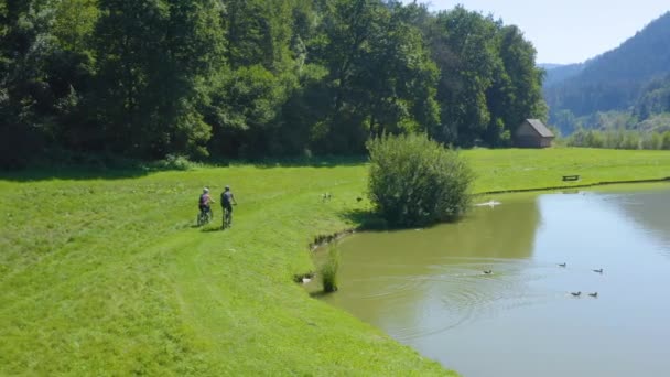 男と女のカップルは 彼らが通過すると 水に飛んでアヒルと湖の周りに自転車に乗って 空中ドローンの右のショットを明らかに スロベニアウォドニ公園 — ストック動画