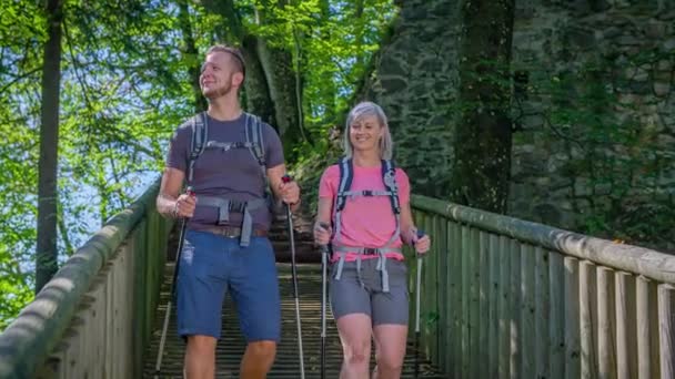 年轻的一对带着背包和北方手杖的运动型夫妇 沿着有树木和砖墙的木制人行道走下去 — 图库视频影像
