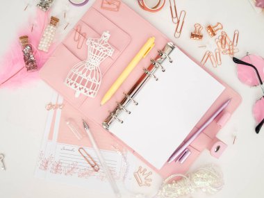 Şirin kırtasiyeli pembe bir planlayıcının üst görüntüsü. Beyaz manken figürlü pembe bir planlamacı. Beyaz arkaplan ve güzel aksesuar kalemleri, düğmeleri, broşürleri olan bir planlayıcı.