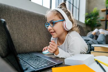 8 yaşında gözlüklü bir kız, odada kanepede uzanan dizüstü bilgisayarla evden internetten ders çalışıyor. Koronavirüs salgını sırasında uzaklık öğrenme.