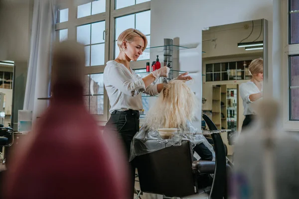 Professional hairdresser dyeing hair of female client at beauty salon in front of a mirror. Selective focus on the hairdresser.