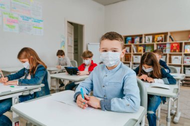 Sınıfta oturan ve kameraya bakan koruyucu maskeli çocuk. İlkokul öğrencileri COVID-19 salgını sırasında ders çalışıyor. Seçici odak.