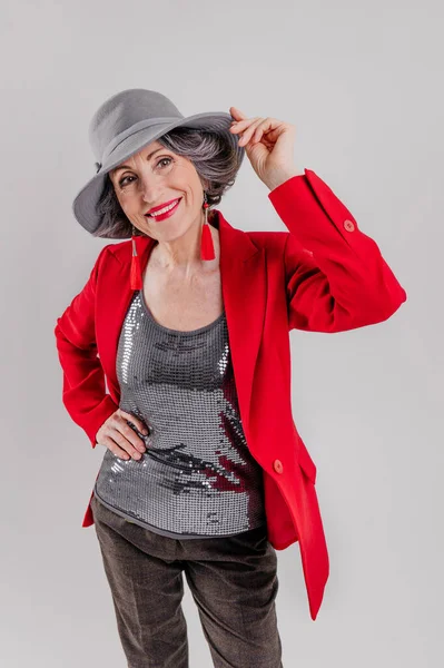 Femme âgée élégante portant un blazer rouge et un chapeau gris sur fond gris — Photo