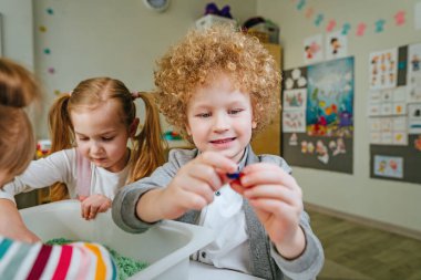 Anaokulunda ya da kreşte oyun öğrenme. Anaokulu öğrencileri, içinde kinetik kum olan bir kabın içinde ahşap boncuklar arıyor ve onları renklerine göre sıralıyorlar. Eğitim konsepti. Seçici odak.