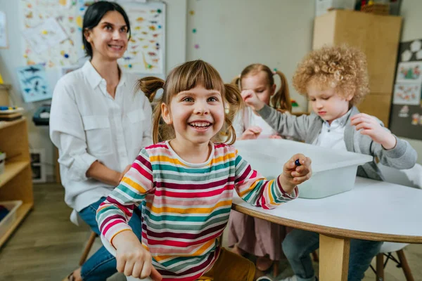 Anaokulunda ya da kreşte oyun öğrenme. Anaokulu öğrencileri, içinde kinetik kum olan bir kabın içinde ahşap boncuklar arıyor ve onları renklerine göre sıralıyorlar. Eğitim konsepti. Seçici odak.