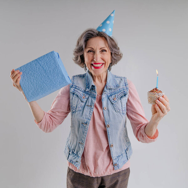 Счастливая пожилая женщина с тортом на день рождения со свечой в шляпе на сером фоне с пробелом для текста.