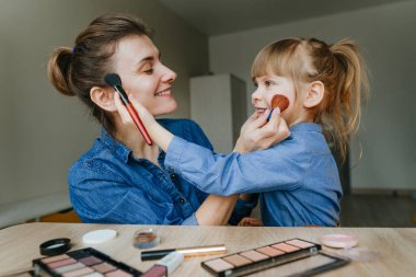 Kapalı alanda makyaj fırçalarıyla birbirlerini tamamlayan genç anne ve küçük kızının portresi. Aile eğlencesi ve güzellik konsepti. Seçici odak.