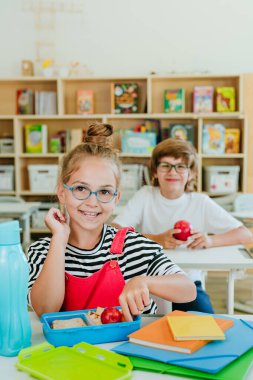 İlkokul öğrencileri, sınıftaki beslenme çantalarından yemek yiyorlar.