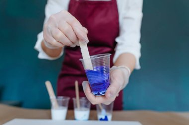 Plastik bardaktaki akrilik boyaları akışkan dökme tekniğiyle boyamak için hazırlayan kadın ressamın yakın çekimi. Seçici odak.
