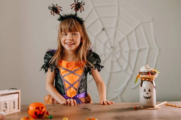 小女孩穿着万圣节女巫服装 戴着蜘蛛头带 坐在桌旁 戴着糖果和怪物奶昔或怪胎奶昔的画像 有选择的重点 — 图库照片