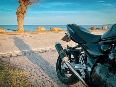 Siyah bir motosiklet denizin kenarında gün ışığında duruyor ve gökyüzü mavi.
